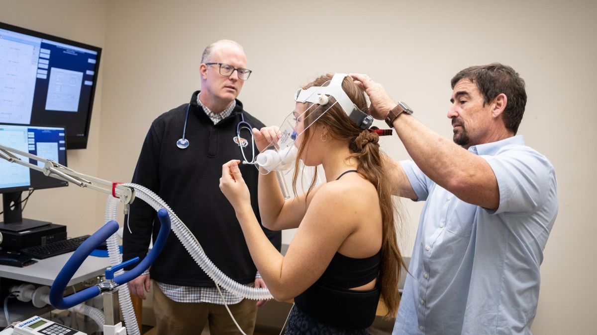 two professors stand next to a student hooked up to a machine for exercise testing