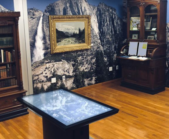 a room with an antique desk and bookcase filled with old books, a painting of the Sierra Nevadas, and a digital screen for viewing information