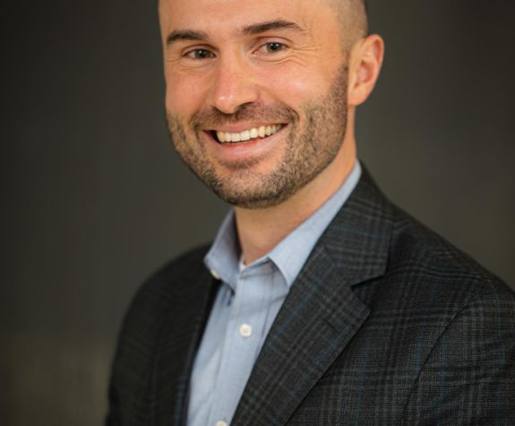 A portrait of a man wearing a suit in front of a gray background