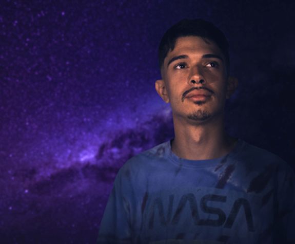a student standing in front of a purple-colored space background looks up toward the sky
