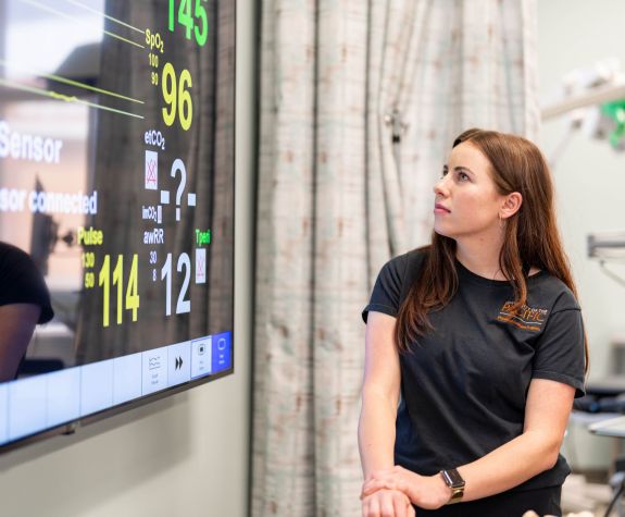 A PA student is shown looking up at a patient monitor. 