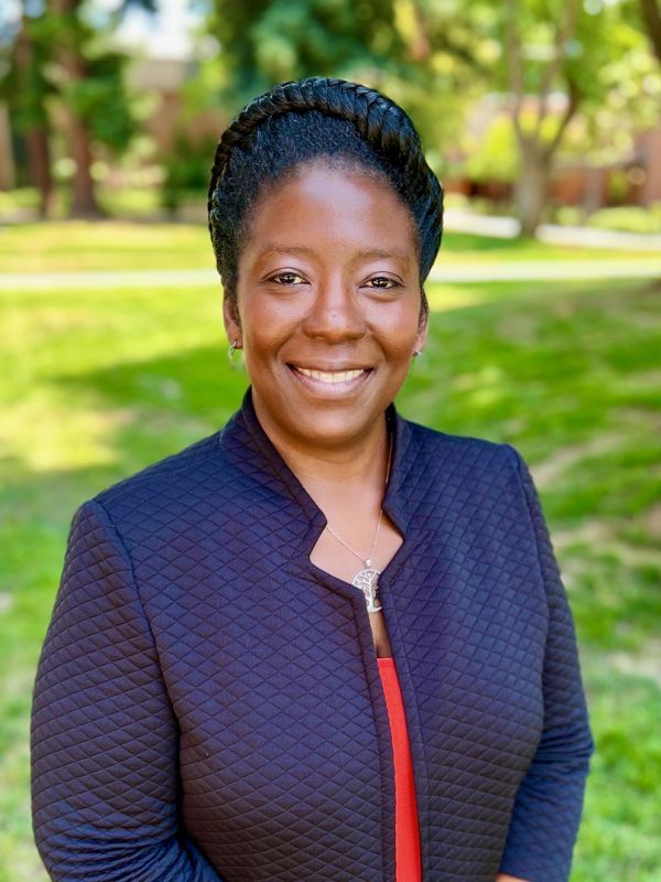 The photo shows the smiling face of Dr. Tanisha Sparks. she is wearing a dark blue jacked over an orange shirt while standing in the shade of a tree on the quad at the Sacramento campus.