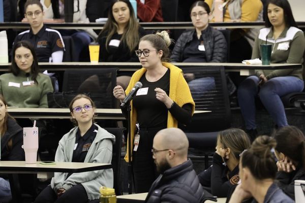 Photo shows a student standing and asking a question at a microphone. She is surrounded by other students who are all seated. 
