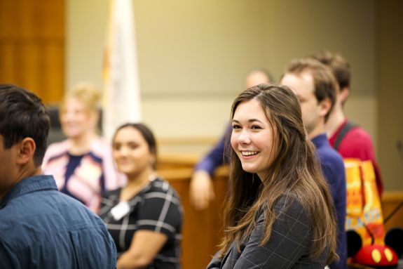 A woman stands in a courtroom among other law students.