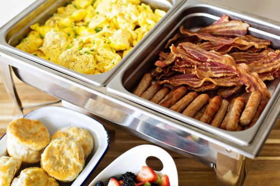An image of eggs, bacon, fresh fruit and breakfast rolls
