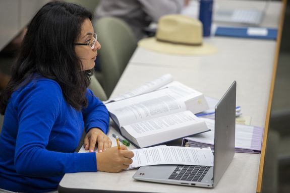 A woman sits behind a laptop with books and notes surrounding her