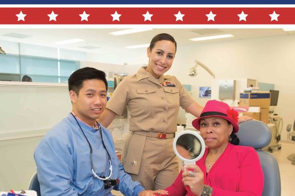 a male dentist with a female member of the military standing and a female patient in the dentist chair holding a mirror
