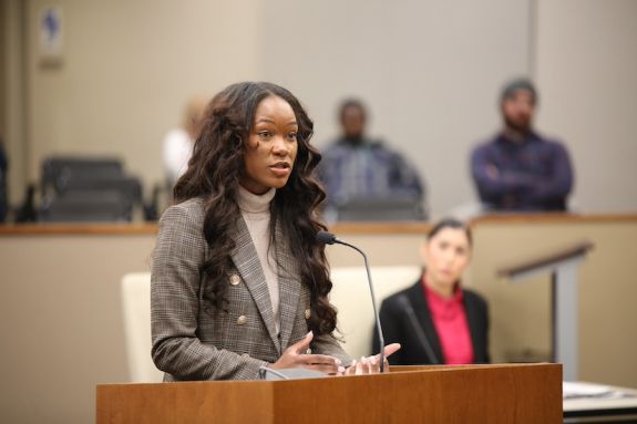Woman in grey blazer speaks at a podium.