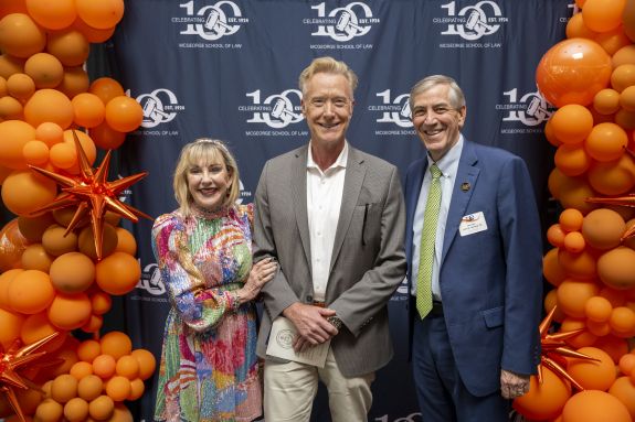 Three people pose for a photo in front of a blue backdrop and orange balloons