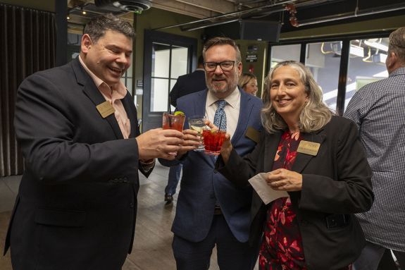 3 people in business attire holding drinks and smiling at the camera. 