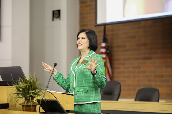 A woman in a green suit jacket specks behind a podium gesturing with her hands at the crowd. 