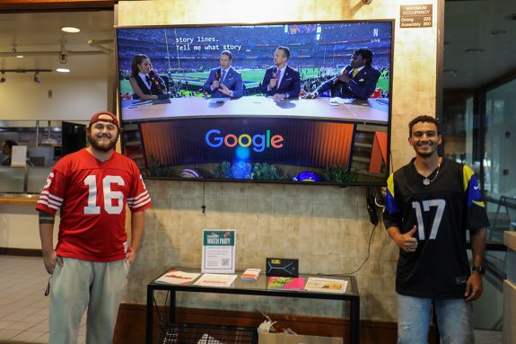 Two men wearing jerseys pose for a photo by a TV