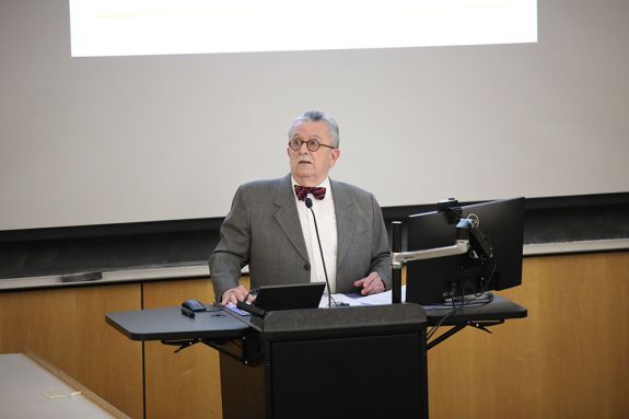 A professor in a grey suit stands in front of a class teaching. 