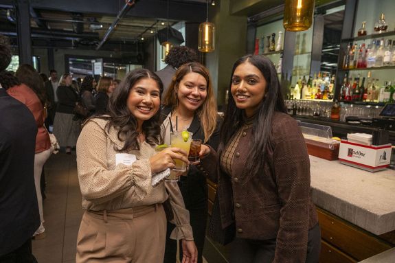Three women pose for a photo holding drinks in celebration.