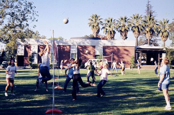 A group of students play a game of volleyball in a large green grass covered quad. 
