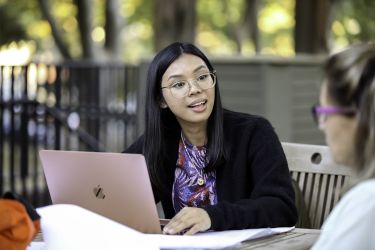 Student sitting in outdoor setting with a laptop, talking to another student. 