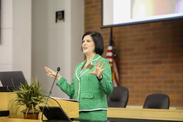 A woman in a green suit jacket specks behind a podium gesturing with her hands at the crowd. 