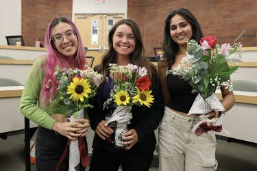 Three women pose and smile for a photo holding bouquets of flowers.