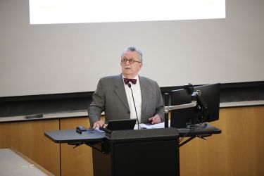 A professor in a grey suit stands in front of a class teaching. 