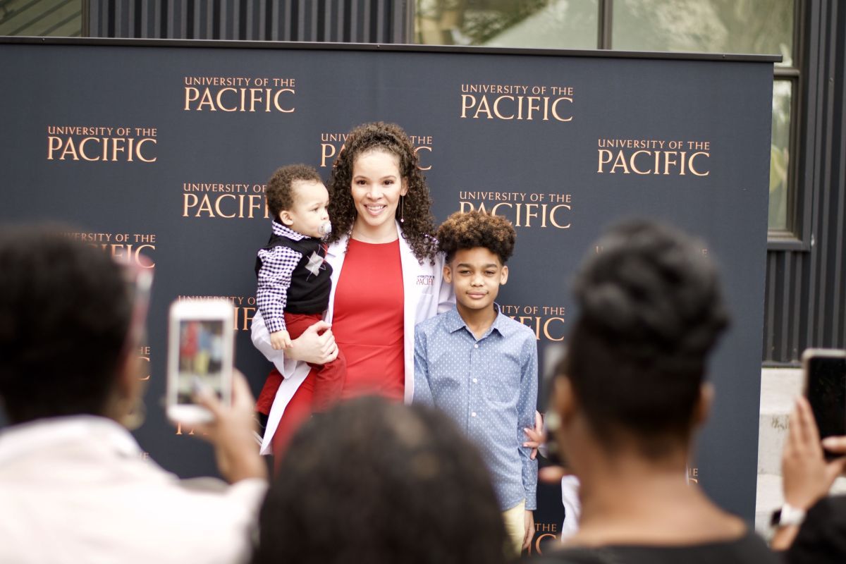 Photo shows a smiling PA student holding her young son while her older son stands beside her as people in the audience snap their photo.