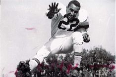 Dick Bass (running back) mid-air with a football clutched under his left arm with his right arm and right leg extended.