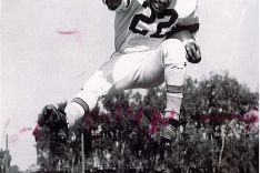 Dick Bass (running back) mid-air with a football clutched under his left arm with his right arm and right leg extended.