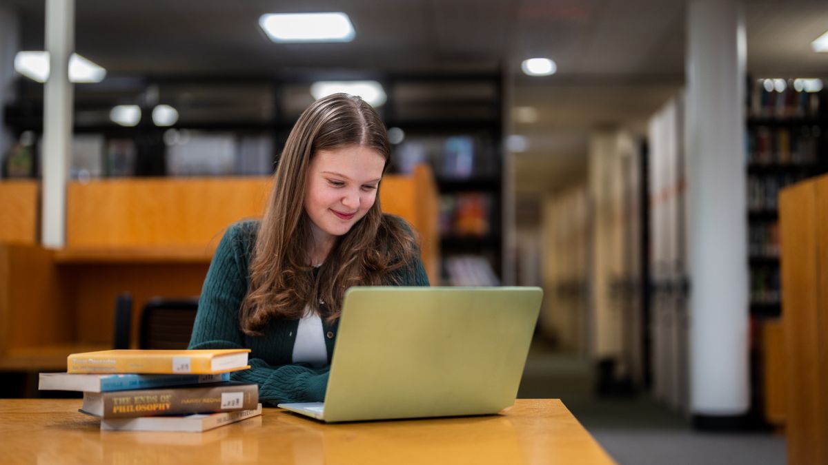 a student sits at a table looking at a laptop