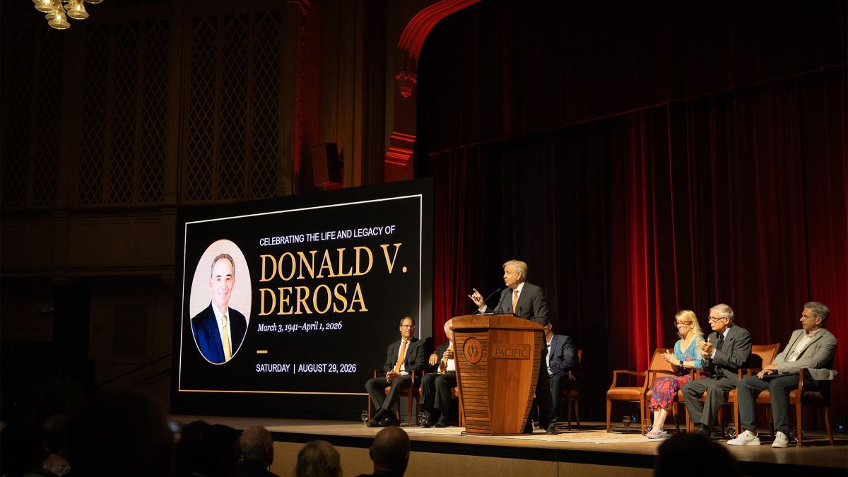 several people sit on a stage next to a large screen that reads "Celebrating the Life and Legacy of Donald V. DeRosa"