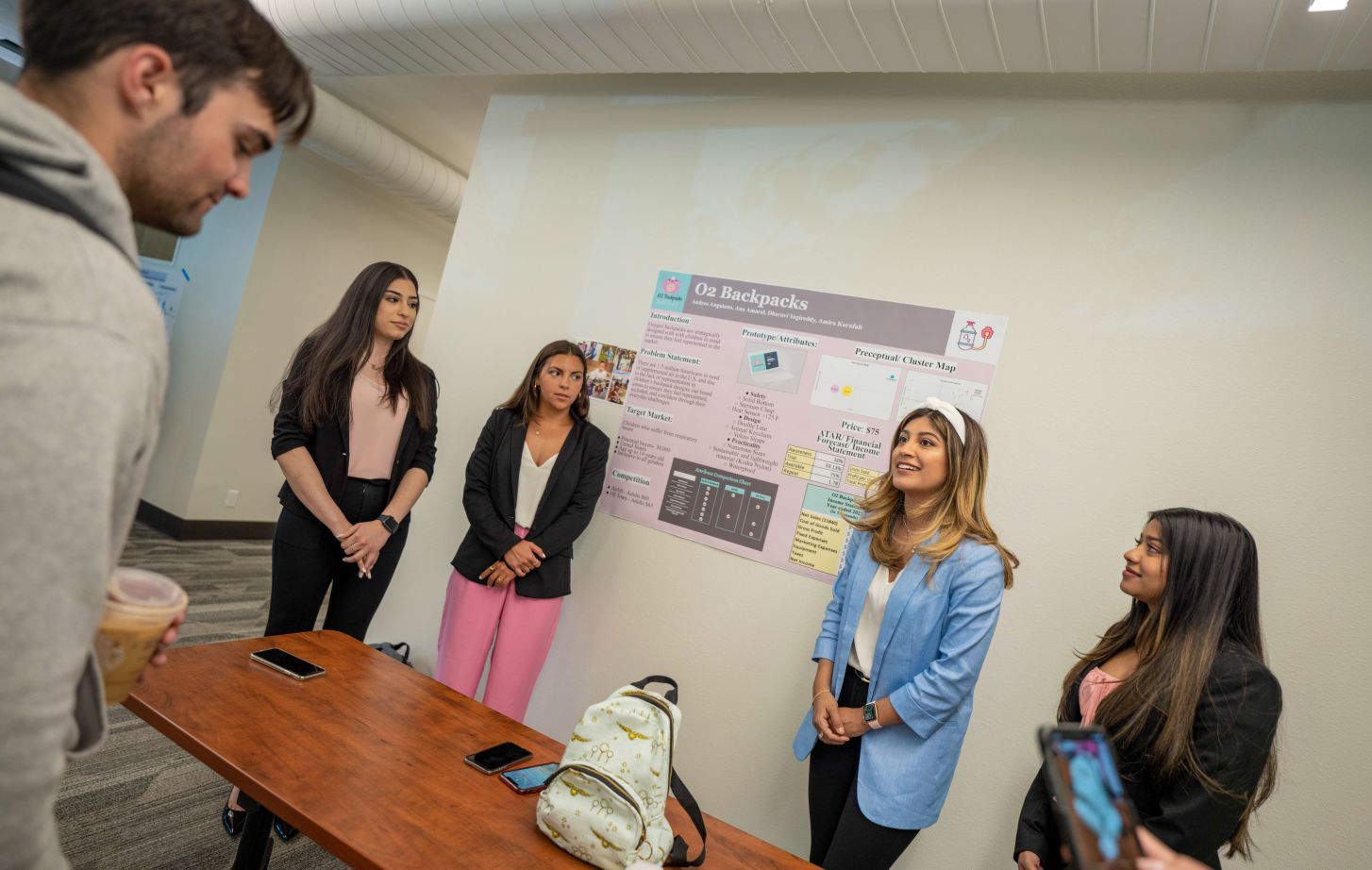 Students stand next to a poster with a backpack on a table in front of them