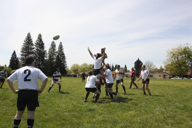 Approximately 10 people in white uniforms play a game of rugby on a grass field.
