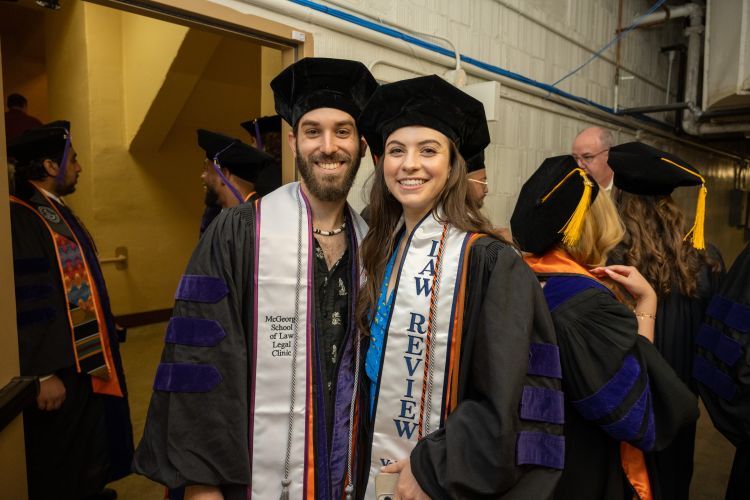 One male individual and one female individual wearing graduation regalia pose for a photo in the basement of an event venue