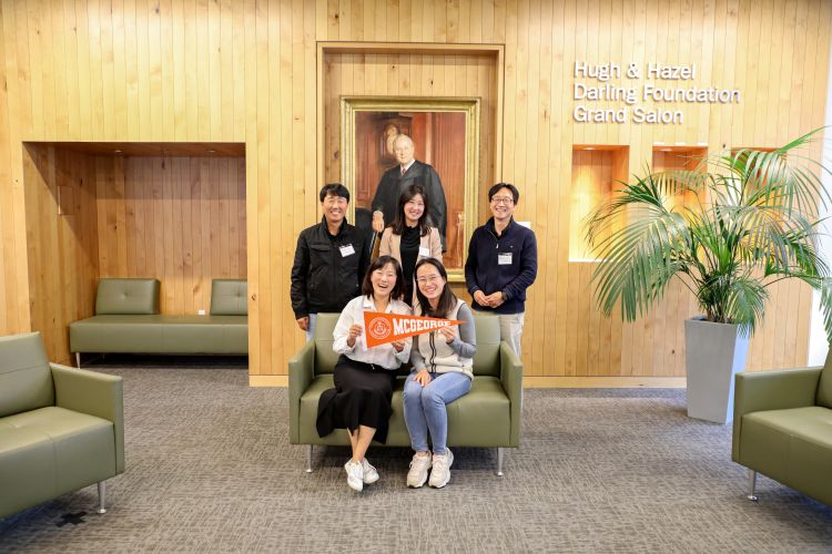 Five Korean individuals pose for a photo. Three are standing, and two are sitting and holding an orange pennant.