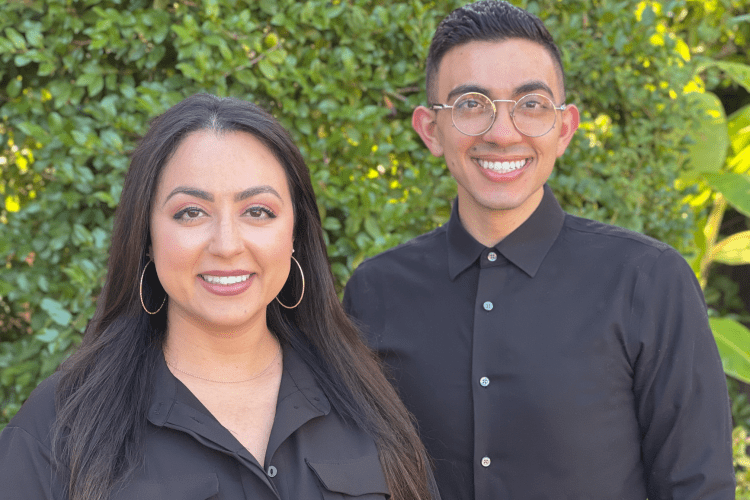Two individuals wearing black pose for a photo in front of shrubbery