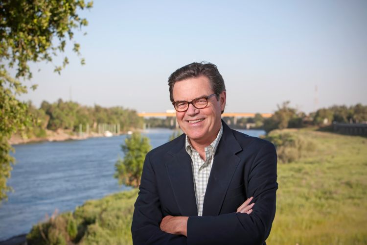 A man in a suit stands smiling in front of a river.