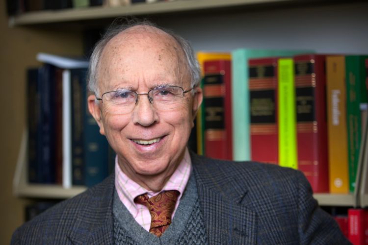 An older gentleman in a suit poses in front of a bookcase.