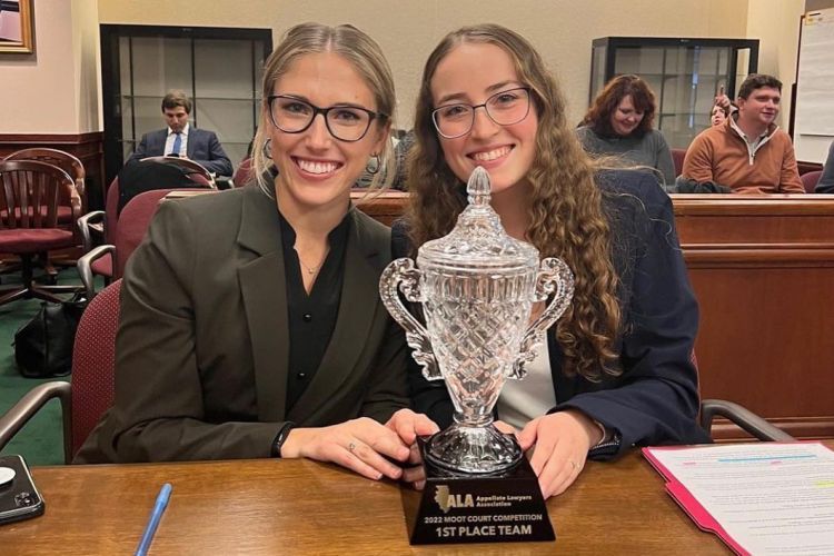 Two female students hold a trophy in a courtroom while seated