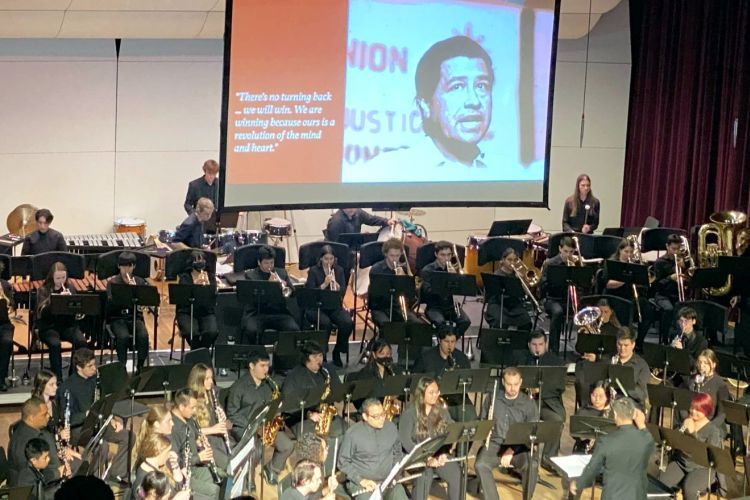 College wind band performing on stage with a photo of Cesar Chaves displayed on a screen