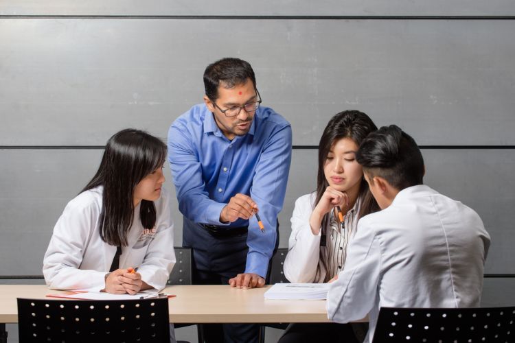 a professor stands next to a table with three seated students