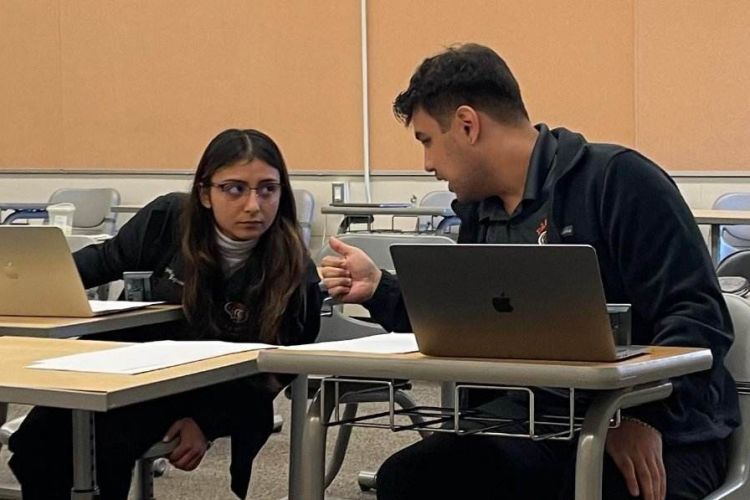 two students sit at desks while talking to each other