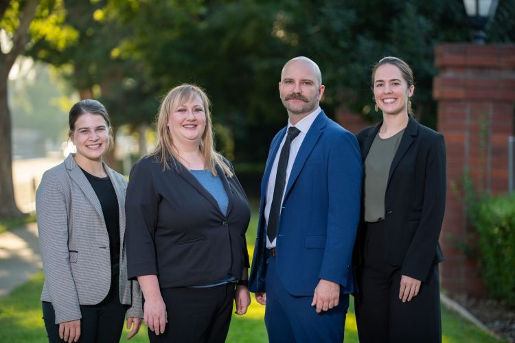 Four people wearing suits pose for a photo outdoors