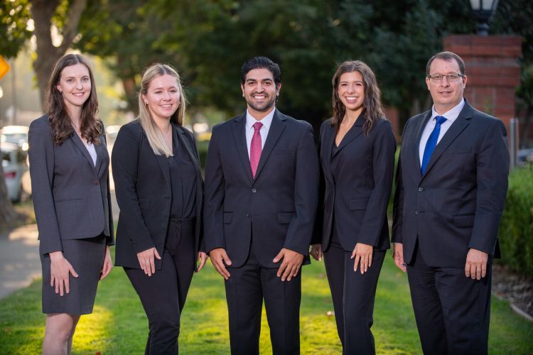 Five people wearing suits pose for a photo outdoors