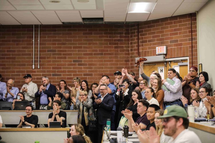 A large group of people stands and claps in a classroom setting.