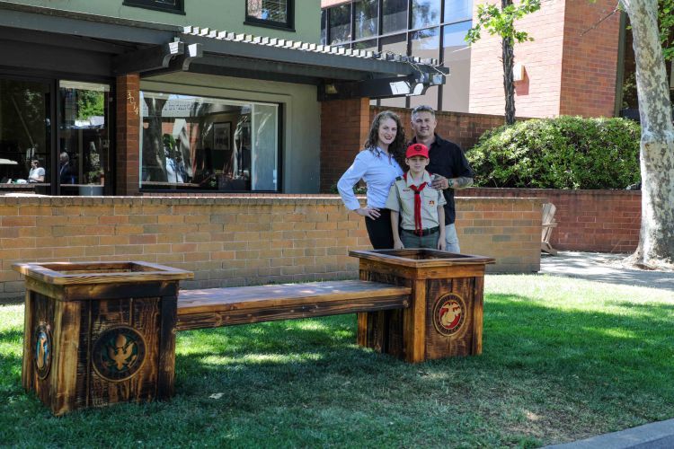 A family of three stands behind a wooden bench with two planters on either side. The bench is located on a grassy area in front of a brick building. The planters have military emblems carved into them.