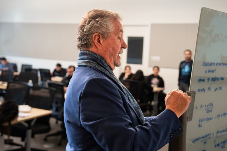 A man writes on a white board in front of a class