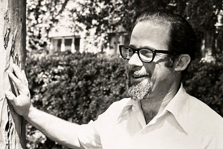 A man wearing glasses stands next to a tree