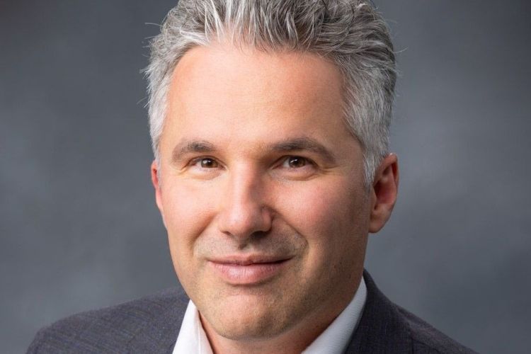 A professional portrait of a man wearing a grey suit against a dark background. 