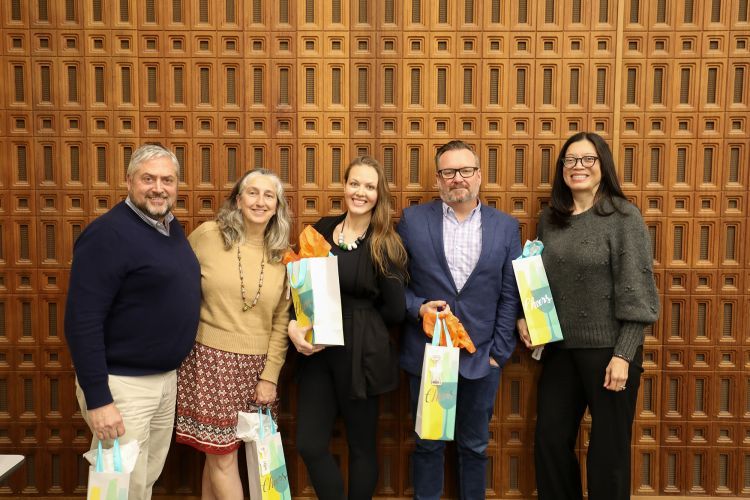 Five people holding gift bags stand in front of a wooden wall smiling for a picture. 