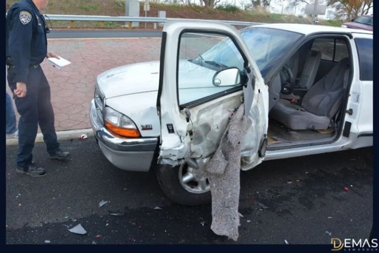 A crushed white truck sits alongside a road with an officer standing off to the stand with a notepad in hand. 
