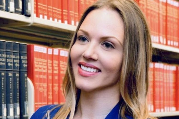 A woman in a blue dress stands in front of a row of law books for a professional photo. 
