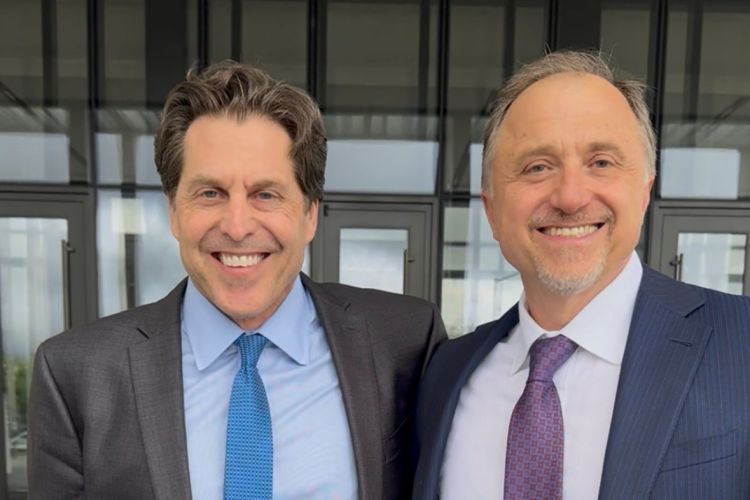 Two men in ties, collared shirts and blazers stand and smile for a photo. 